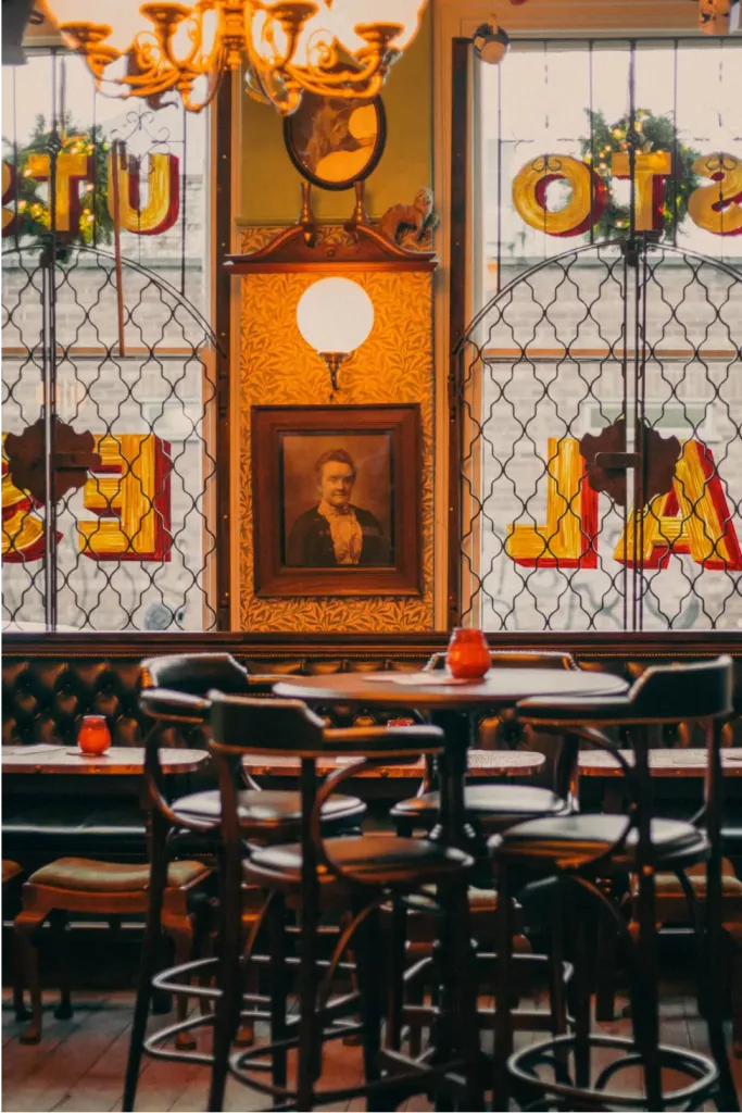 Warm ornate pub interior with high round tables and stools, tufted bench, framed portrait and stained-glass windows.