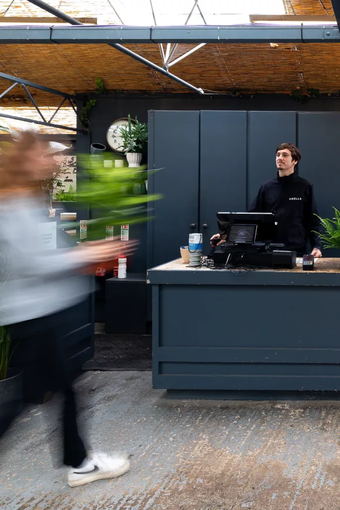 Clerk in black jacket stands behind a shop counter as a blurred customer rushes by carrying a tall green potted plant.