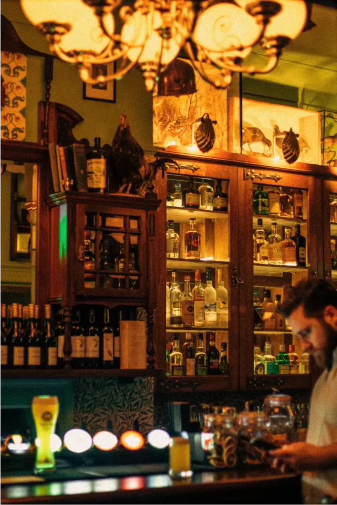 Warmly lit pub interior with a wooden glass-fronted liquor cabinet, a beer on the bar and a bearded man checking his phone.