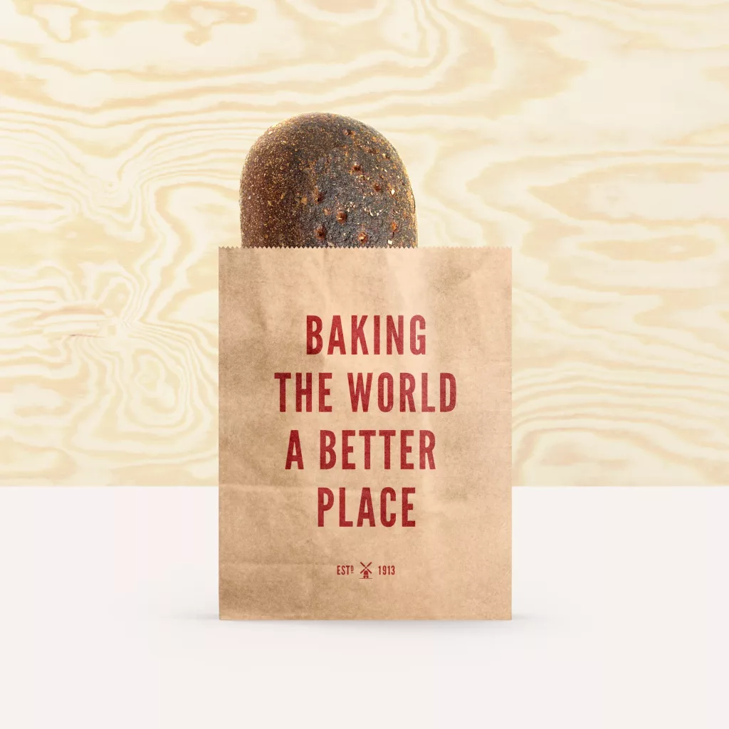 Brown paper bakery bag with a dark seeded loaf poking out and red text 'Baking the world a better place' on the front.