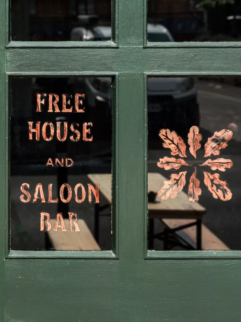 Green-framed window with copper lettering reading FREE HOUSE AND SALOON BAR and a painted leaf motif.