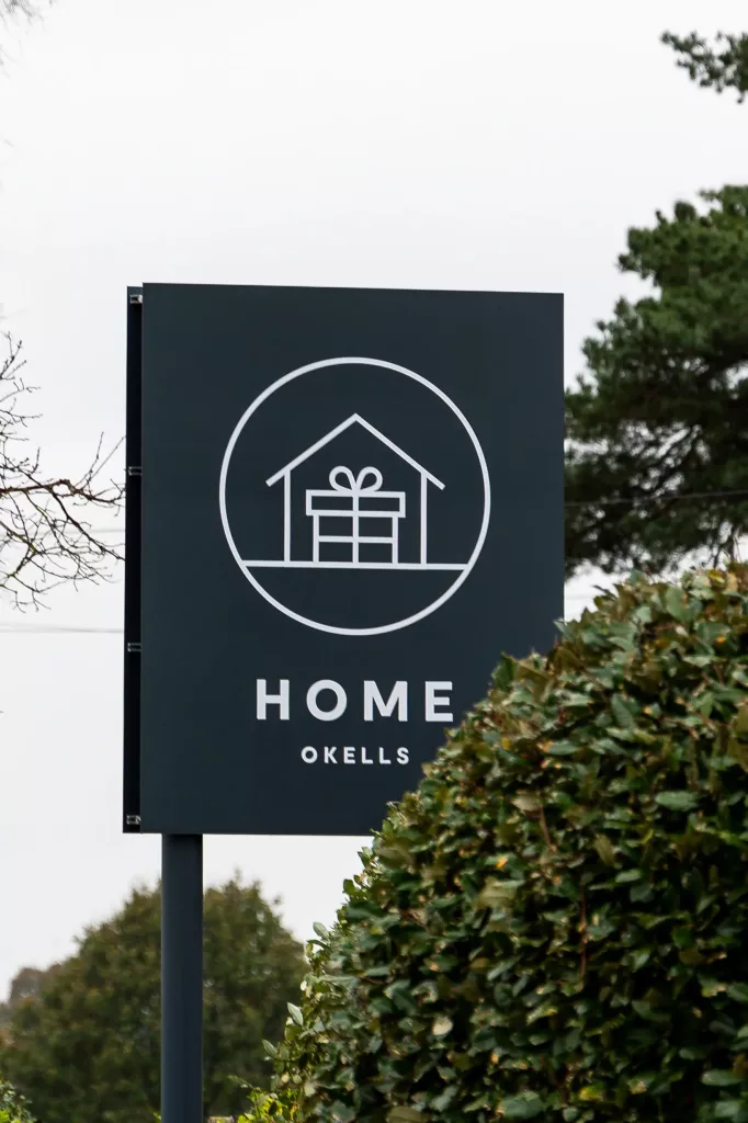 Dark sign on a post featuring a white line-drawn house enclosing a wrapped gift and the text HOME OKELLS, with hedge and trees nearby.