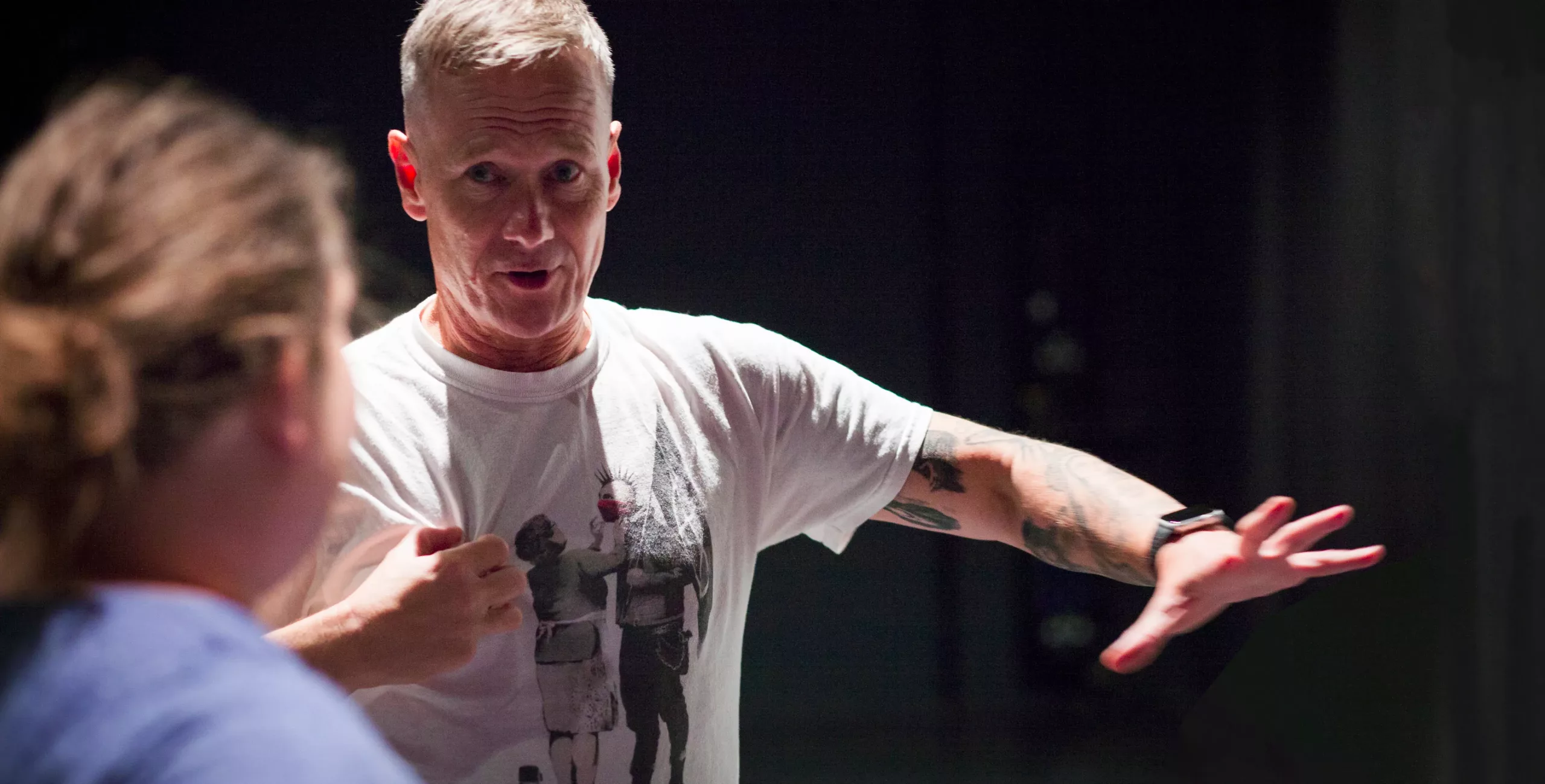 Middle-aged man with short blond hair in a white graphic T-shirt, tattooed arm outstretched, speaking to a blurred person on a dark stage.