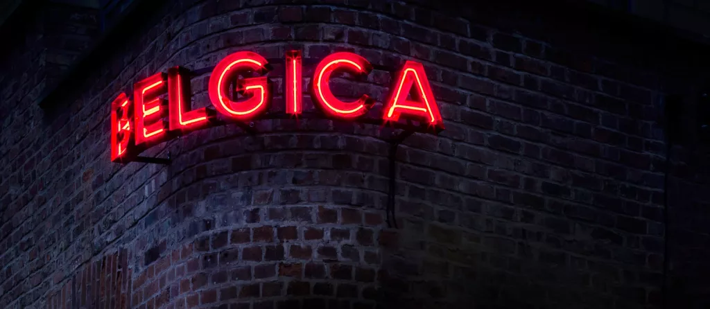 Red neon sign reading BELGICA glowing on the curved corner of a dark brick building at night.