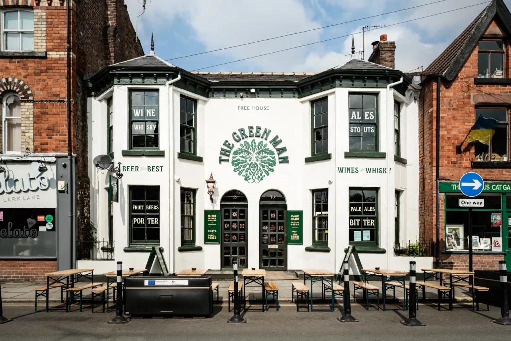 White and green two-storey pub The Green Man with leaf crest, arched double doors and outdoor wooden benches.