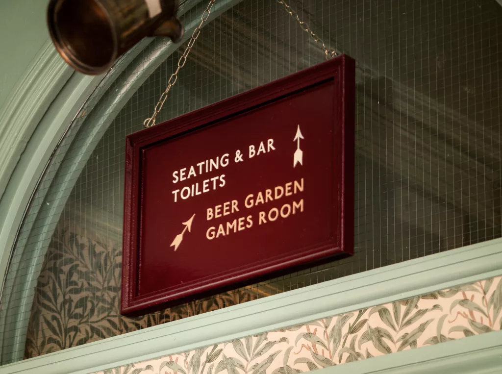 Maroon hanging sign with white arrows listing Seating & Bar, Toilets, Beer Garden and Games Room in a green arched frame