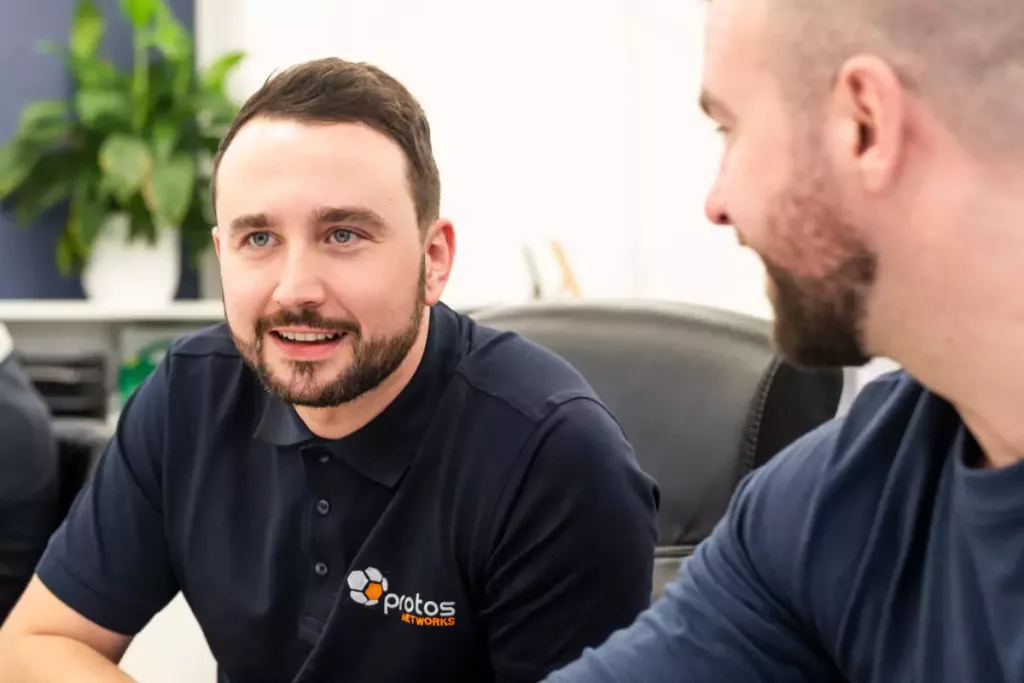 Two men in navy polo shirts talking in an office, one smiling with a Protos Networks logo on his shirt.