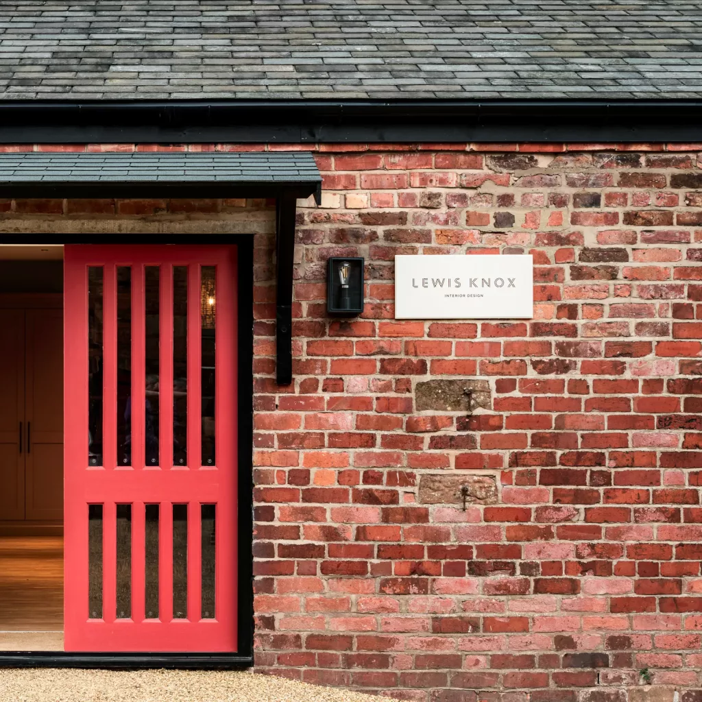 Red slatted door set in weathered red-brick wall beside a white Lewis Knox Interior Design sign and a black wall lantern.