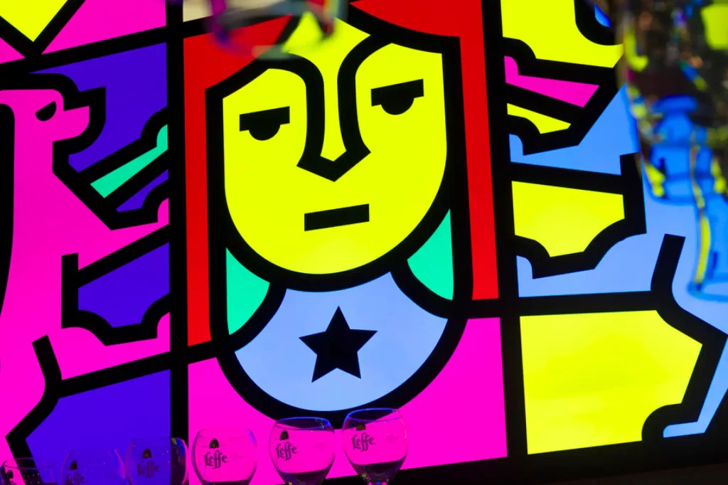 Neon pop-art panel of a yellow-faced figure with red hair, black outlines and a chest star, with Leffe beer glasses at the base.