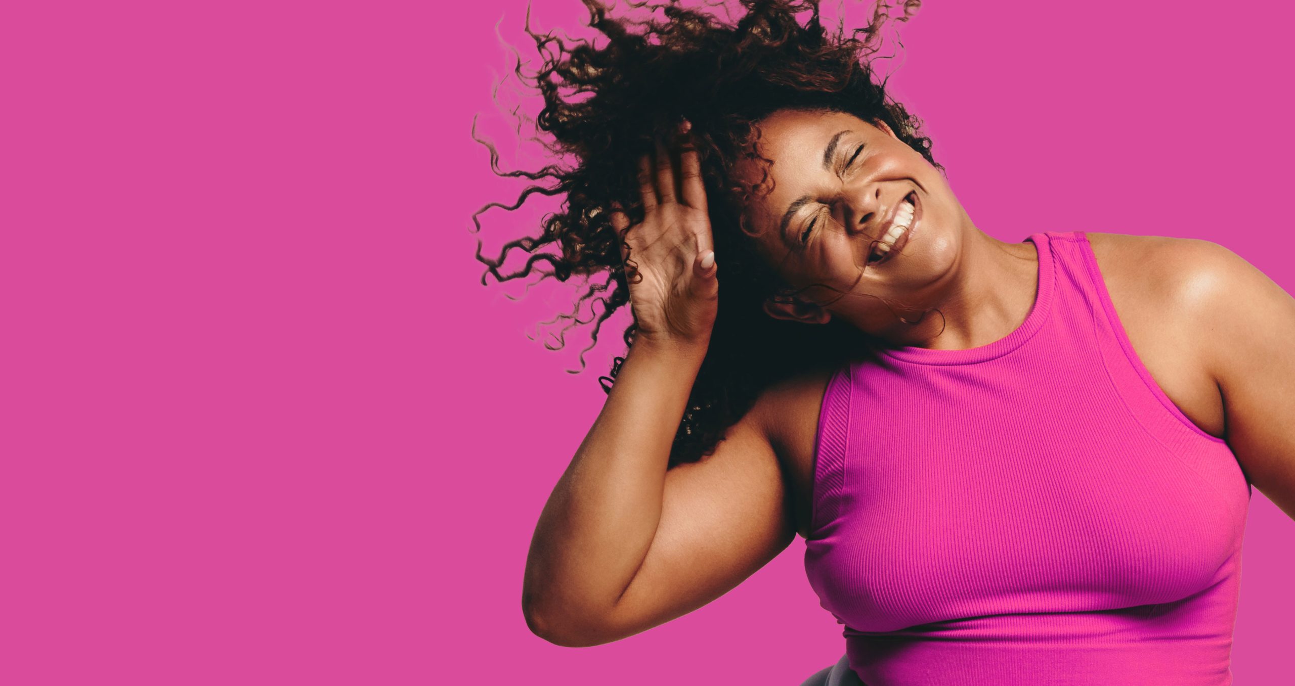 Smiling woman with curly hair tosses it back, wearing a bright pink sleeveless top against a solid magenta background.