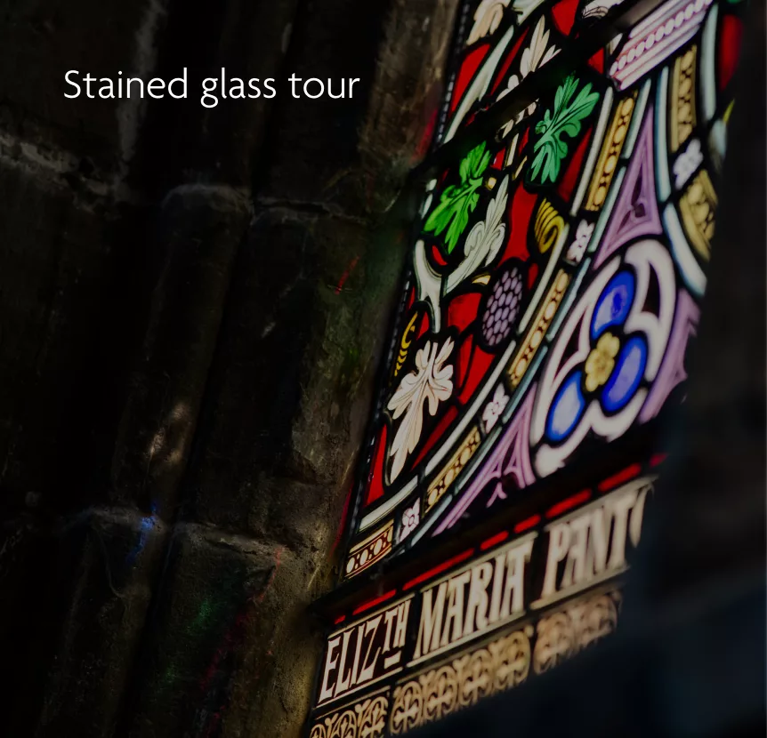 Angled view of a colourful stained-glass window with floral motifs and partial inscription ELIZTH MARIA in a dark stone wall.