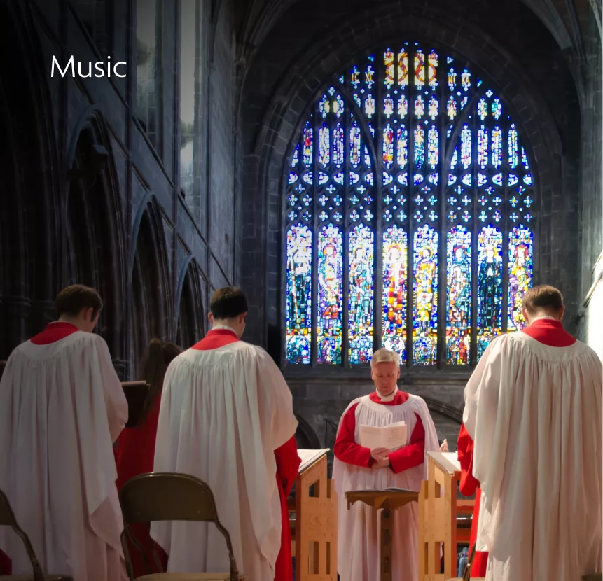 Choir in white robes with red collars singing from sheet music in a cathedral before a large colourful stained-glass window.
