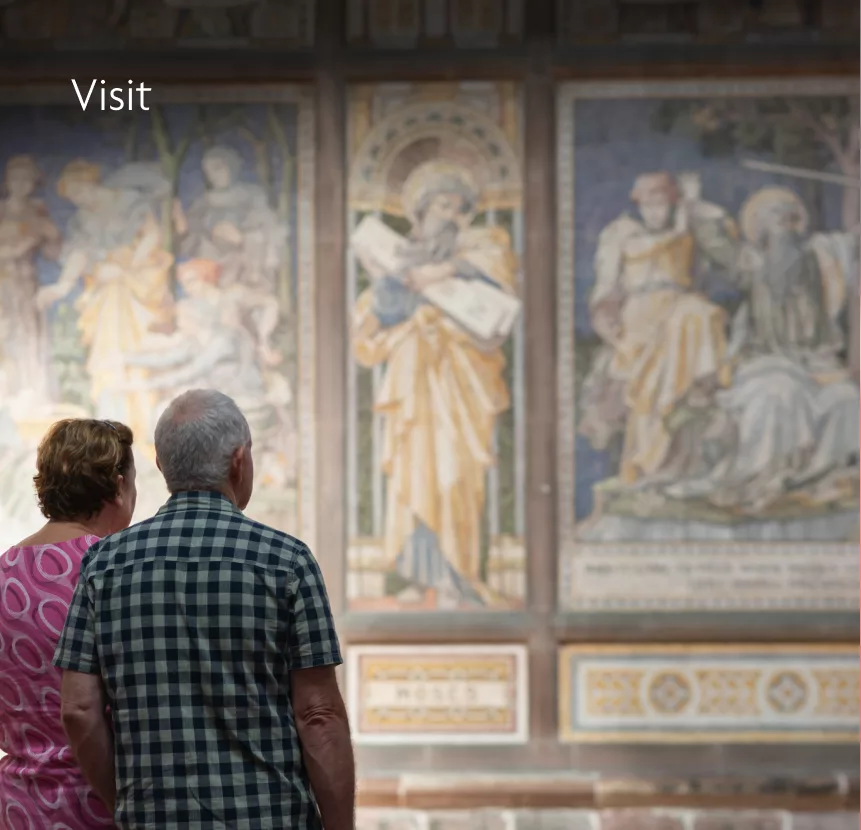 Older couple from behind viewing large religious wall frescoes in a gallery, Visit visible in the top-left.