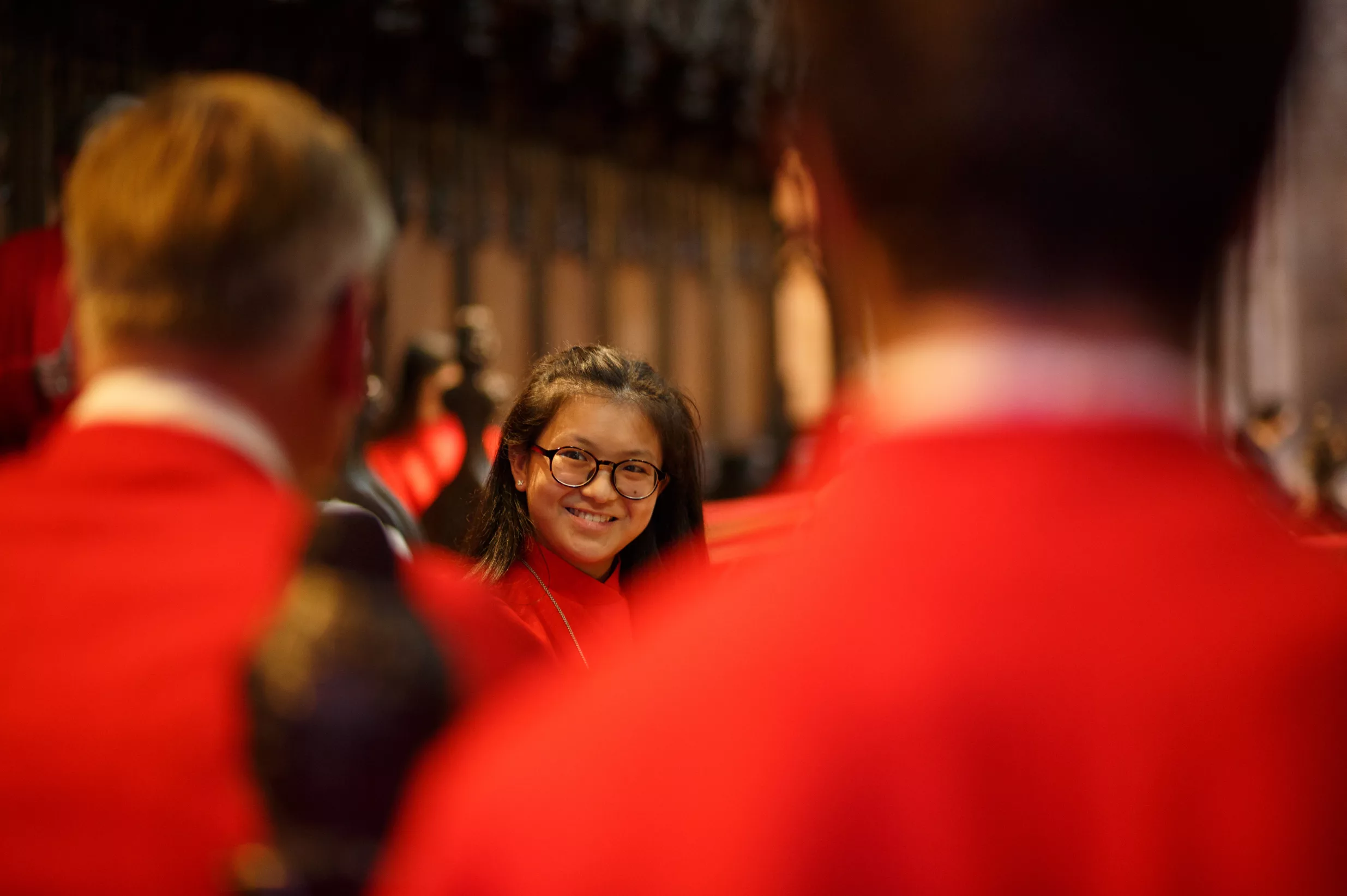 Smiling young woman in glasses and a red choir robe seated among blurred fellow choristers in a church.