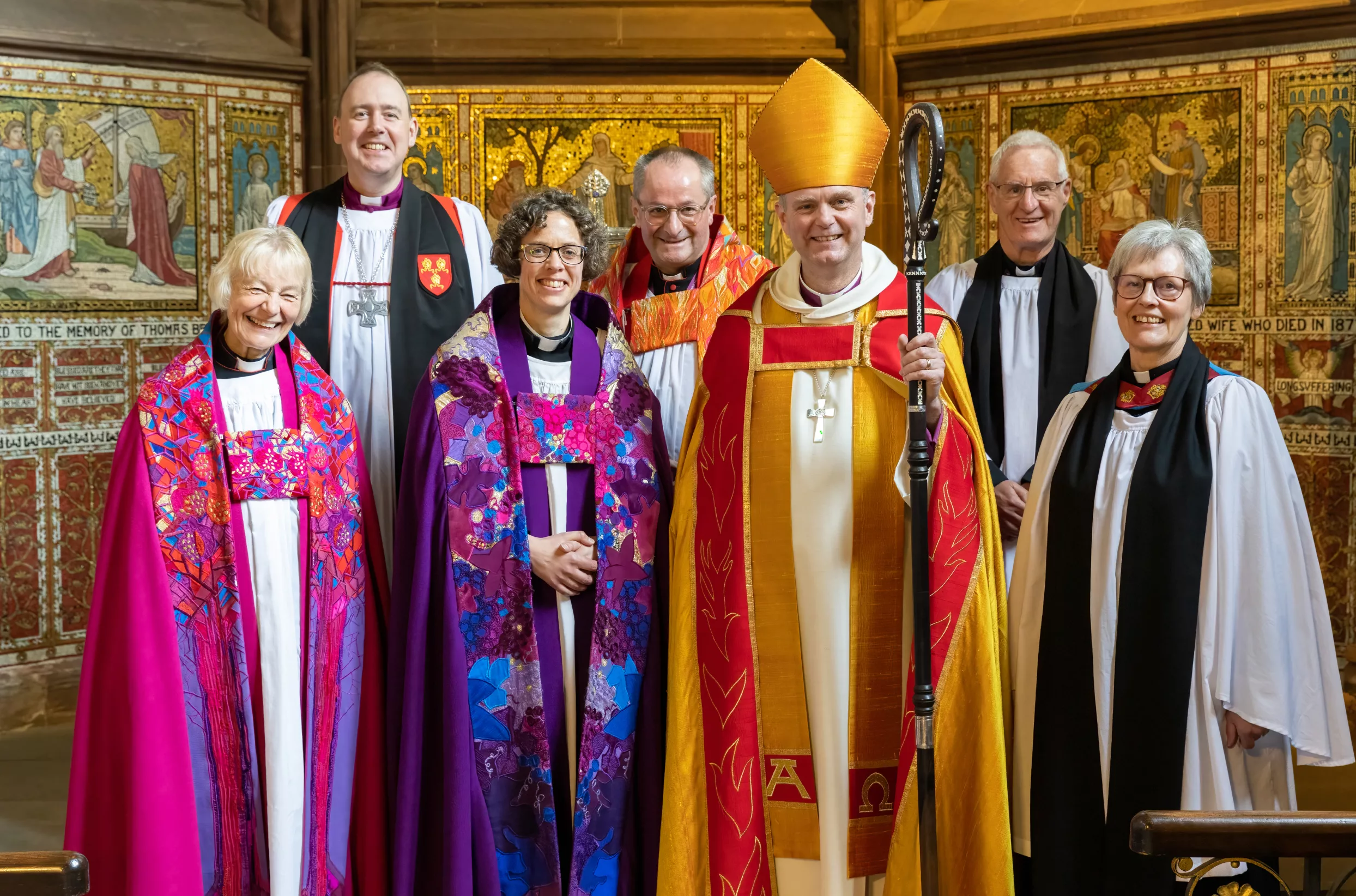Group of clergy in colourful liturgical robes, centre bishop in gold mitre holding a crozier, standing before an ornate gilded chapel wall.