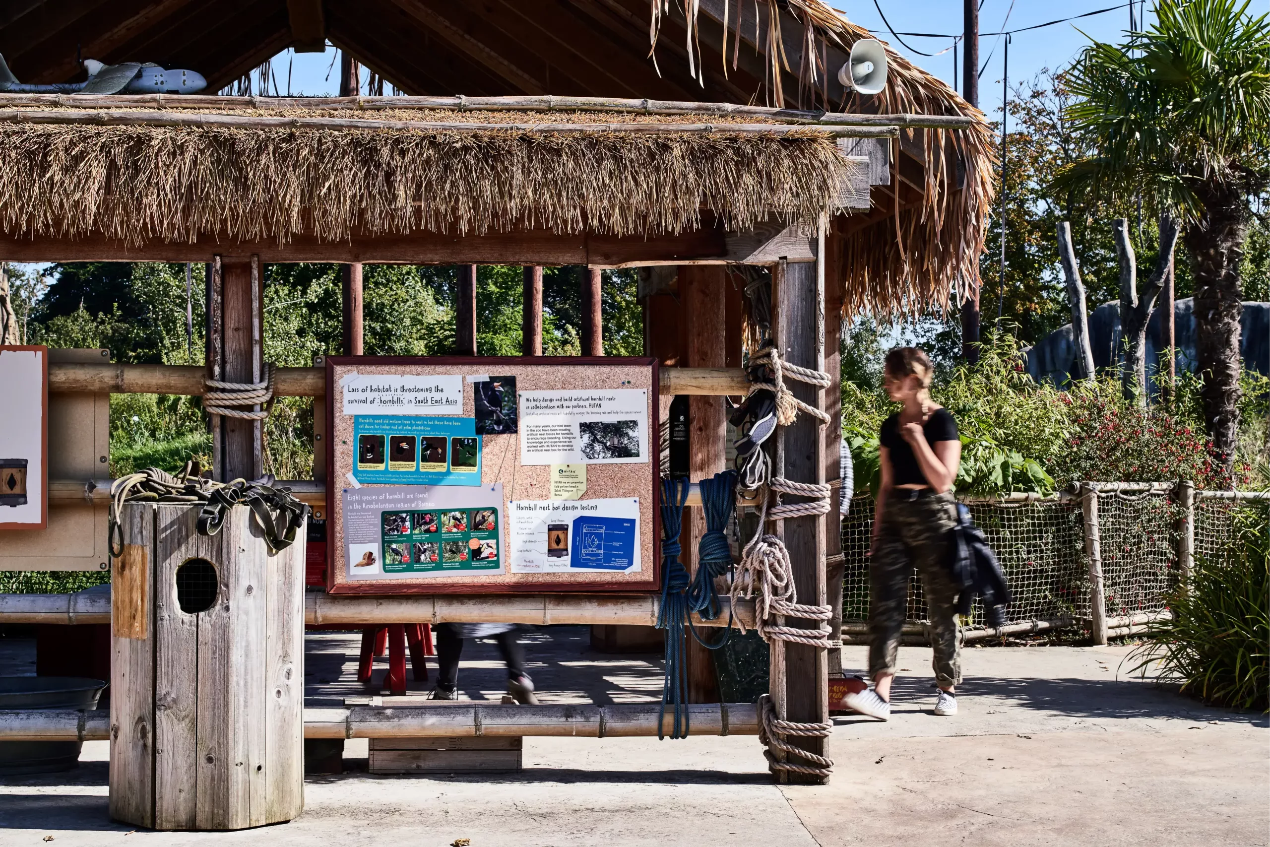 Thatched wooden kiosk with an informational display board and coiled ropes, a woman in camouflage trousers walking past.