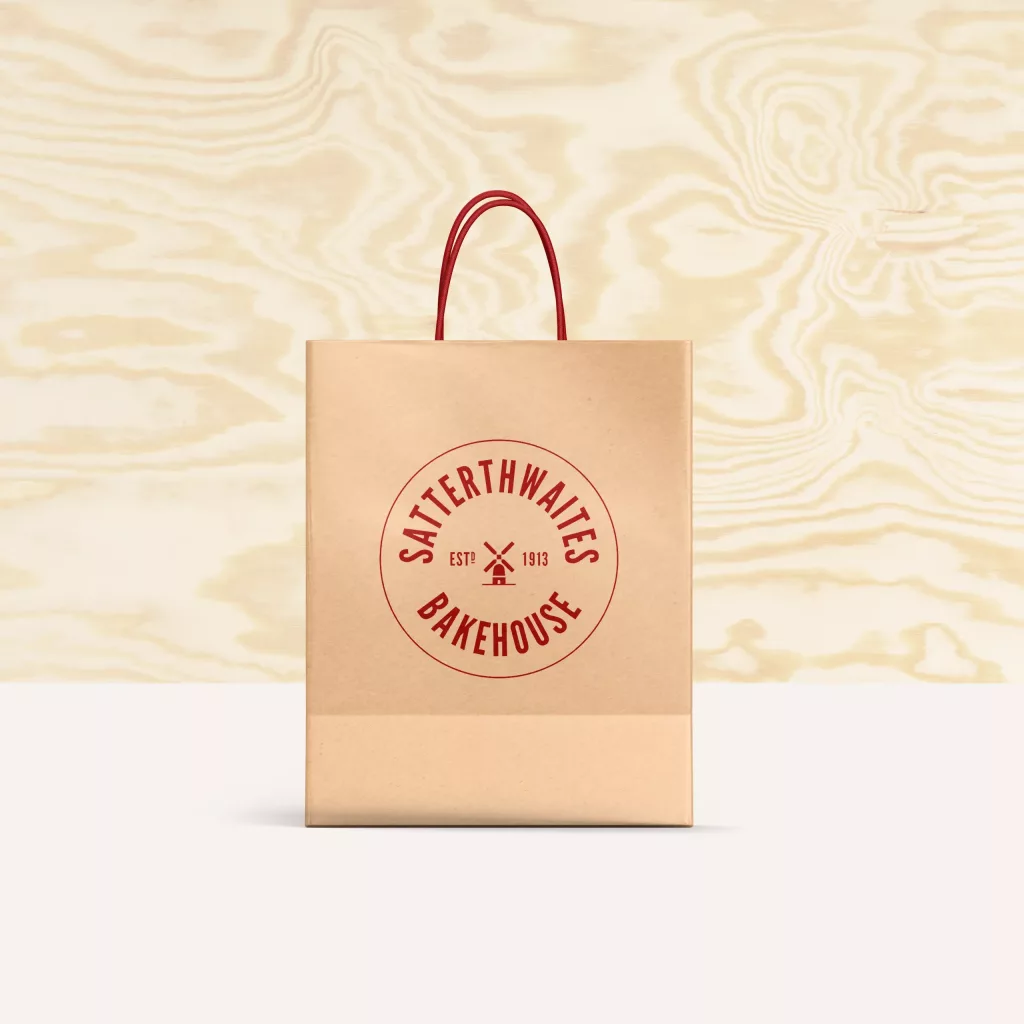 Brown paper bag with red handles and circular red Satterthwaites Bakehouse logo (windmill, Est 1913) on pale wood background.
