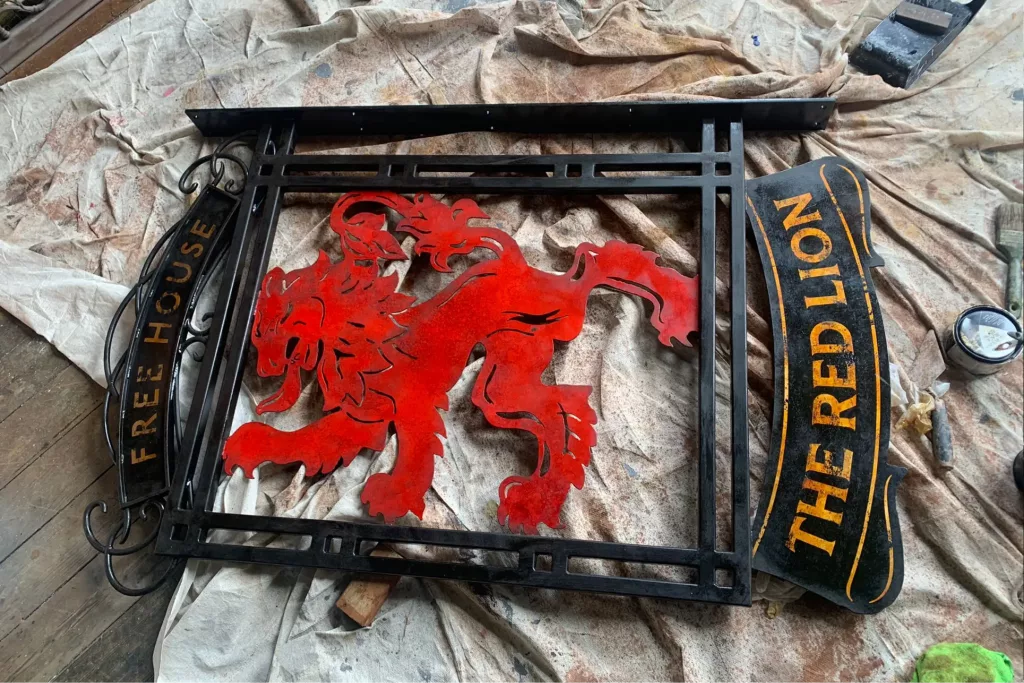 Wrought-iron sign with red lion cutout and plaques reading THE RED LION and FREE HOUSE, on a dusty drop cloth.