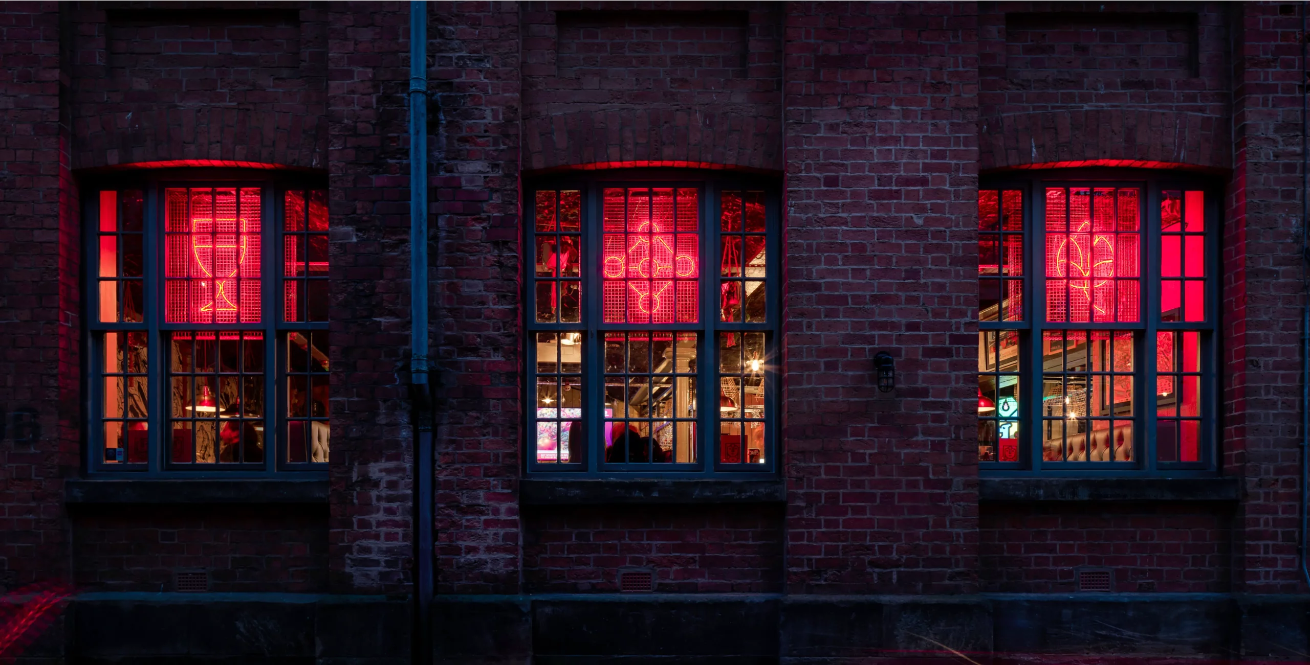 Three tall multi-pane windows set in a red brick wall showing a bar-like interior lit by pink-red neon signs.