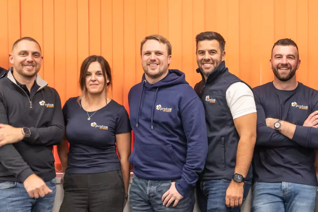 Five smiling colleagues in navy Protos-branded tops standing side by side against orange vertical blinds