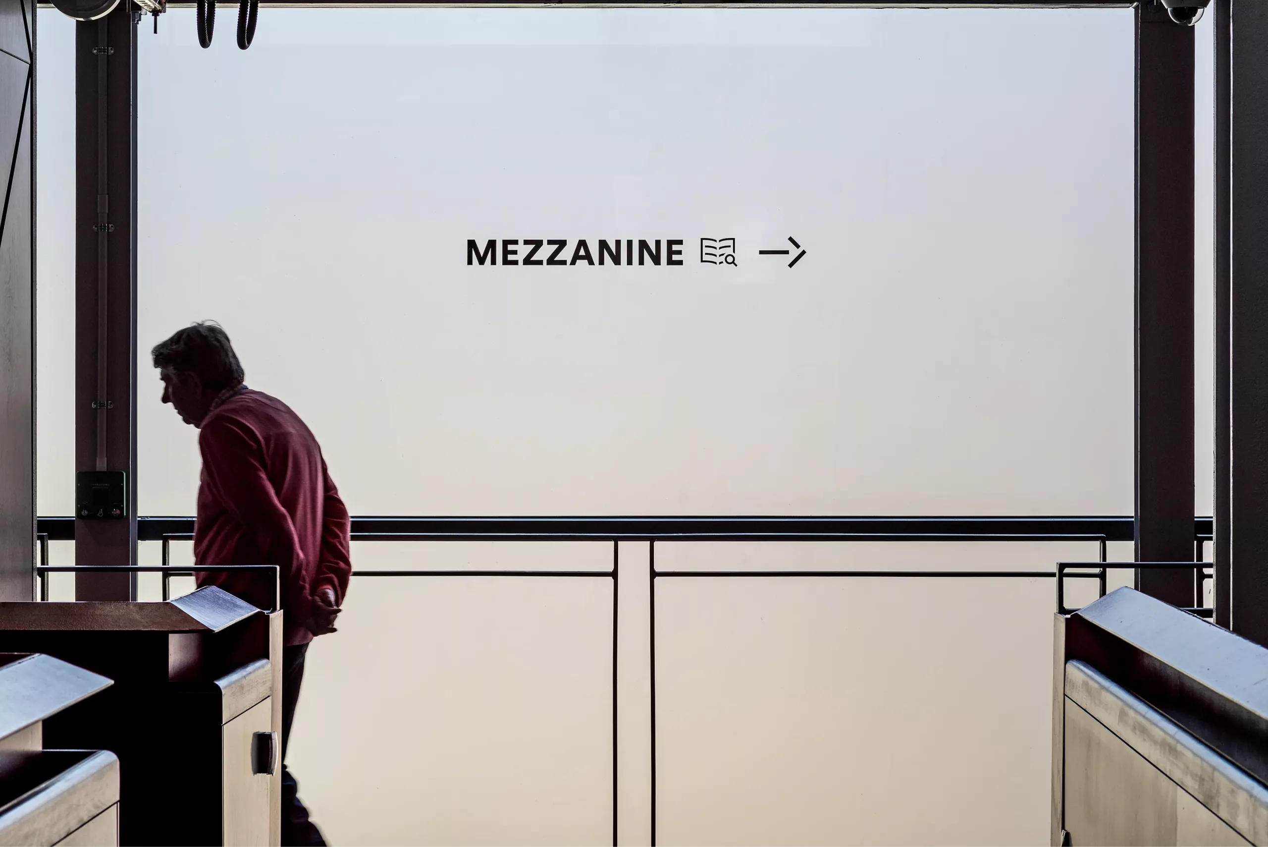 Silhouetted man in a red jacket walking past a railing beneath a MEZZANINE → sign on a pale frosted glass wall.