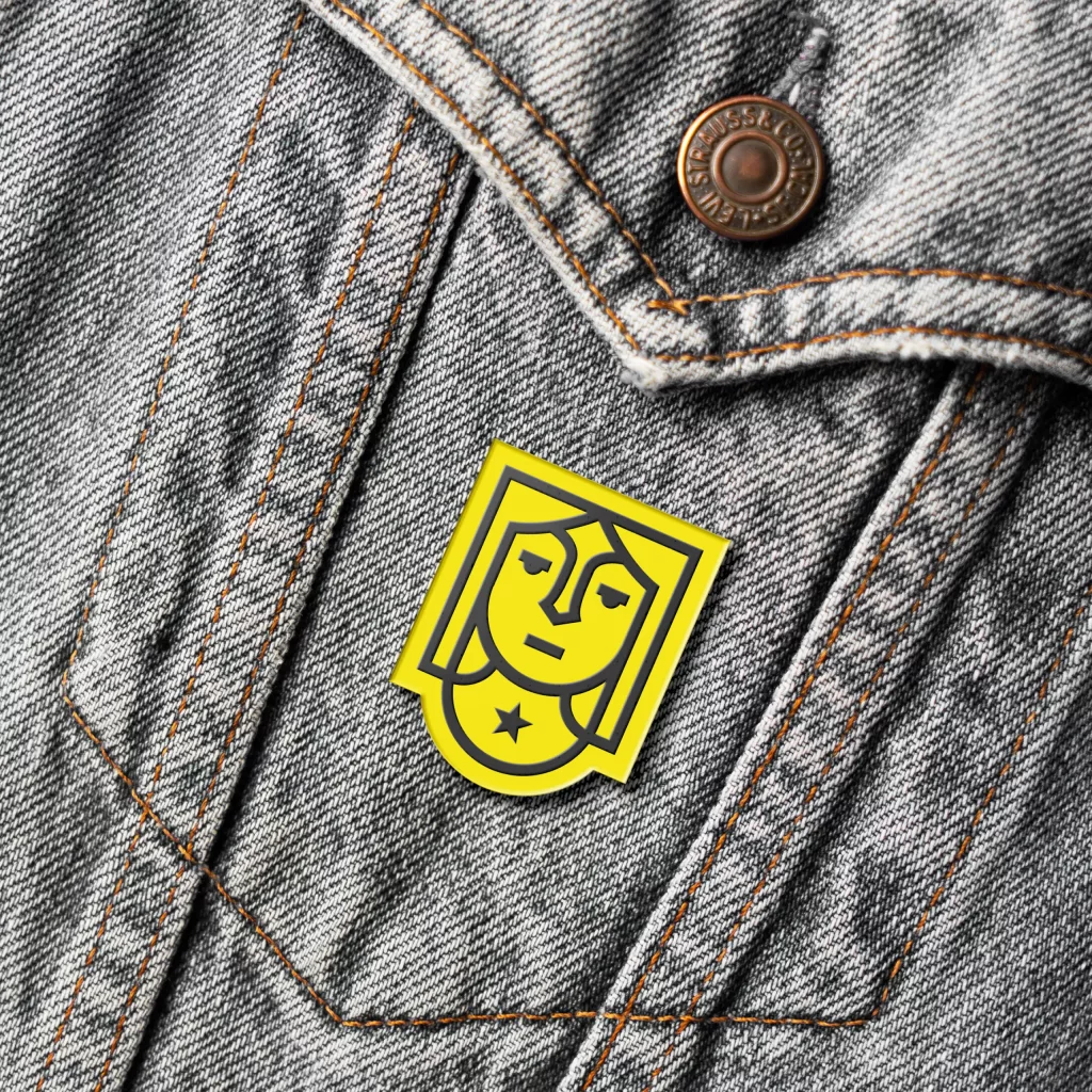 Yellow enamel pin of a stylised face with a star, pinned to grey denim jacket with orange stitching and a brass button.