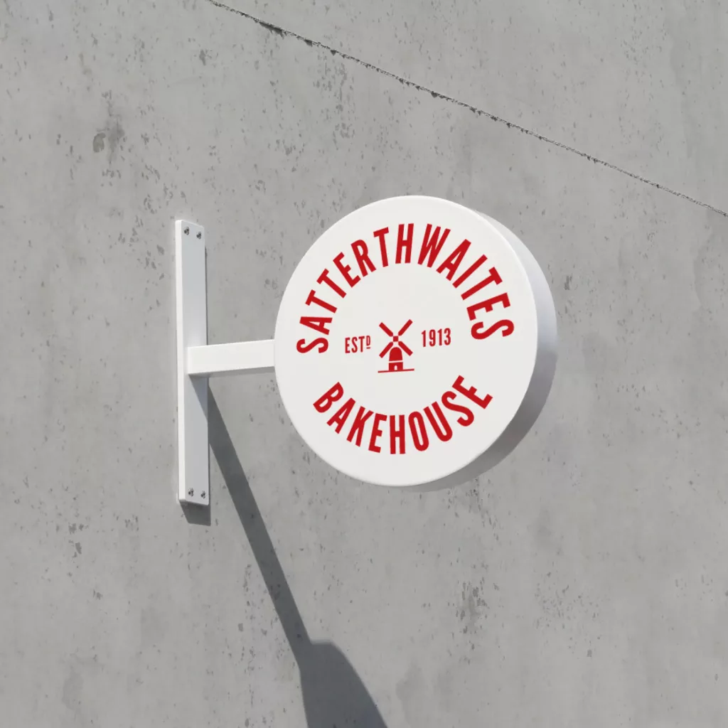 Round white sign reading Satterthwaites Bakehouse in red with windmill logo and Est 1913, mounted on a concrete wall.
