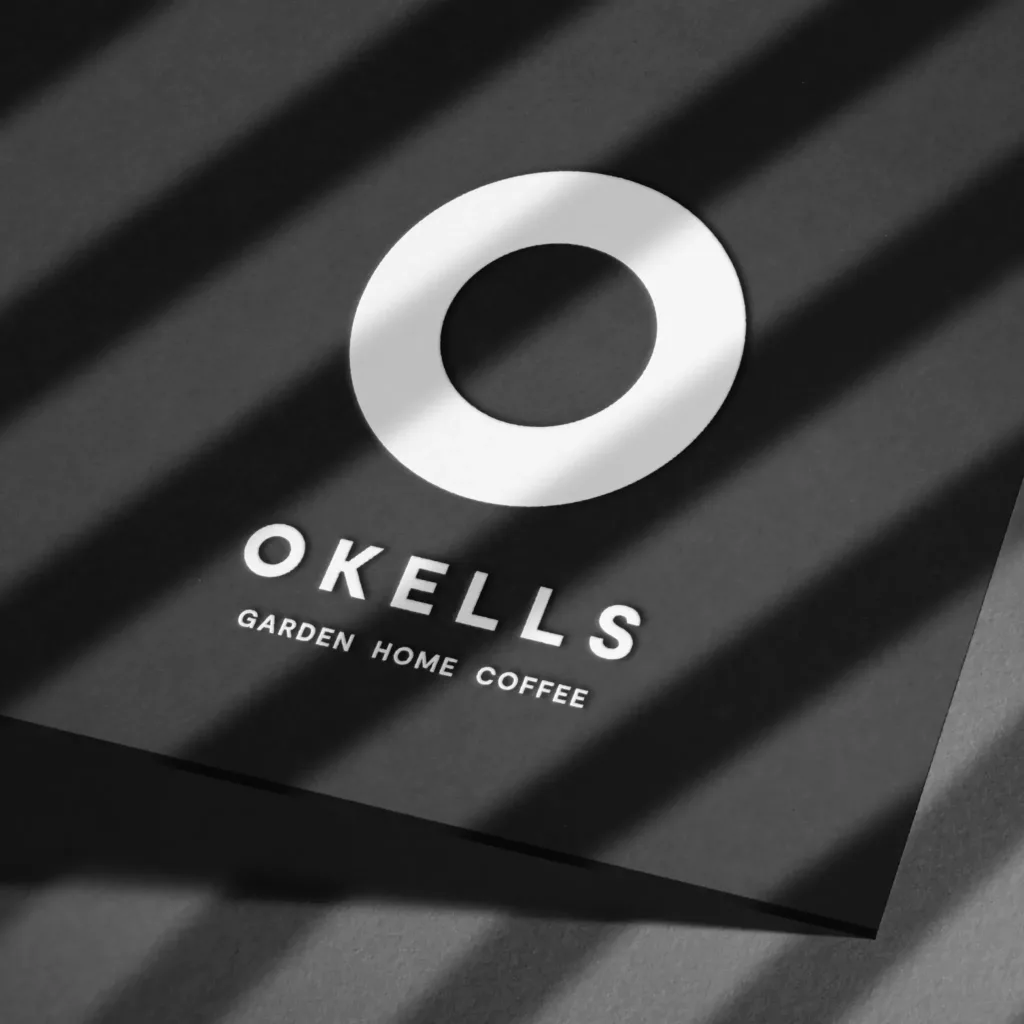 Black card with a white circular logo above OKELLS and the 'Garden Home Coffee' tagline, crossed by diagonal shadows.