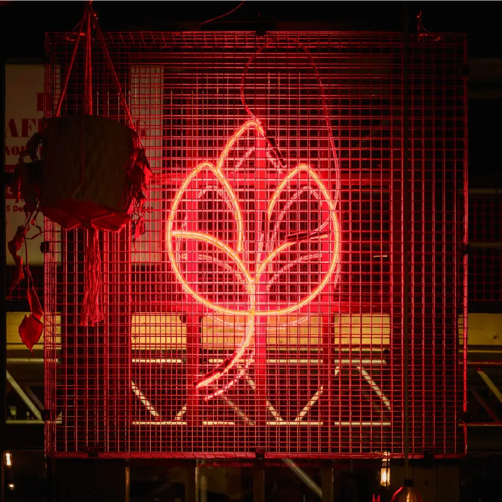 Red neon sign in the shape of a stylised leaf glowing against a metal wire-grid panel, with a hanging planter at left.