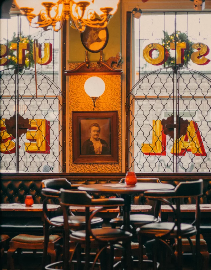 Vintage pub interior with round wooden table and chairs, leather banquette, stained-glass windows and a framed portrait.