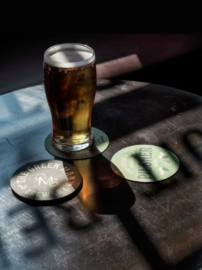 Pint of amber beer with a white head on a dark wooden table beside green brewery coasters, sunlight casting long shadows.