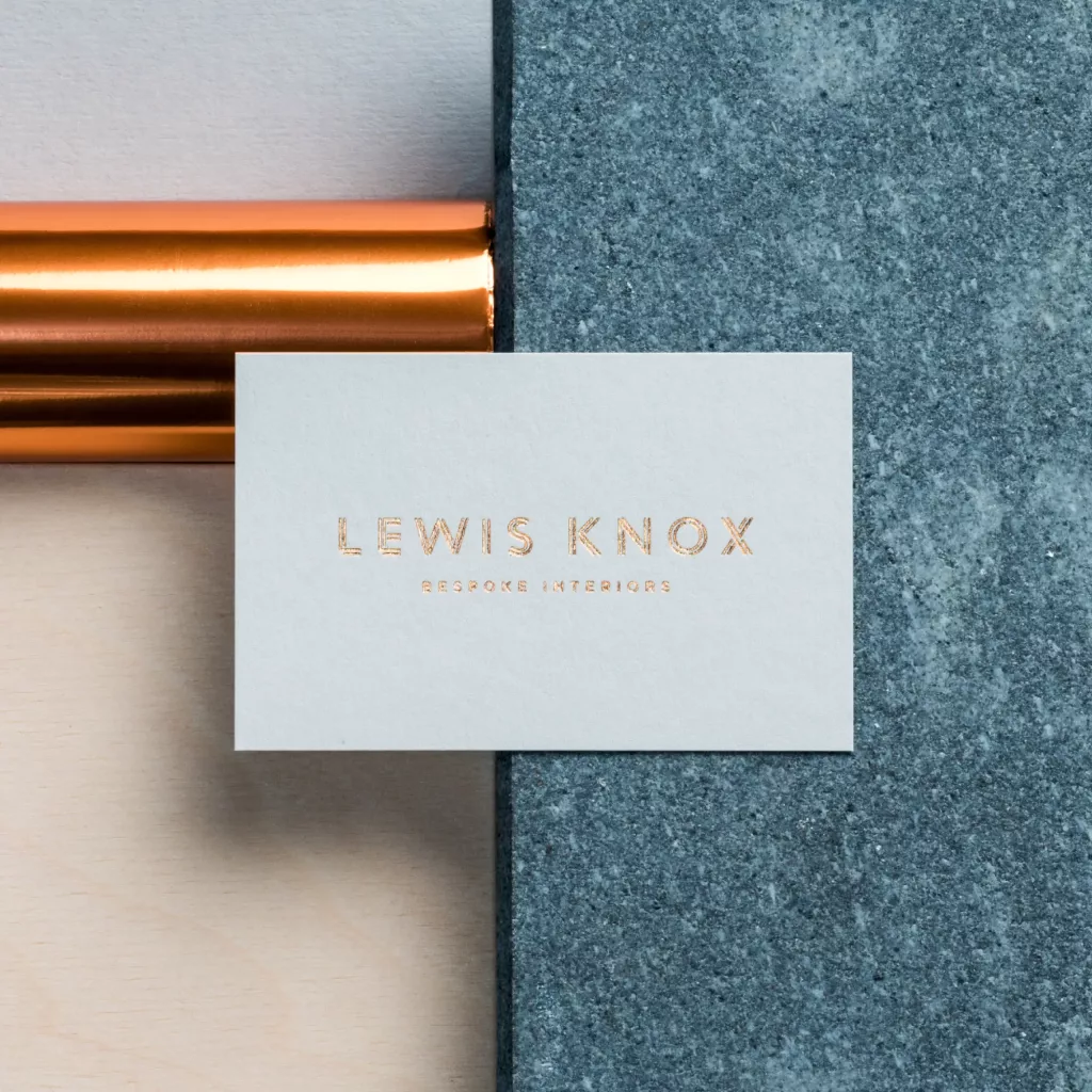 Business card with copper-foil LEWIS KNOX Bespoke Interiors on pale card, set over copper tube, wood and grey stone.