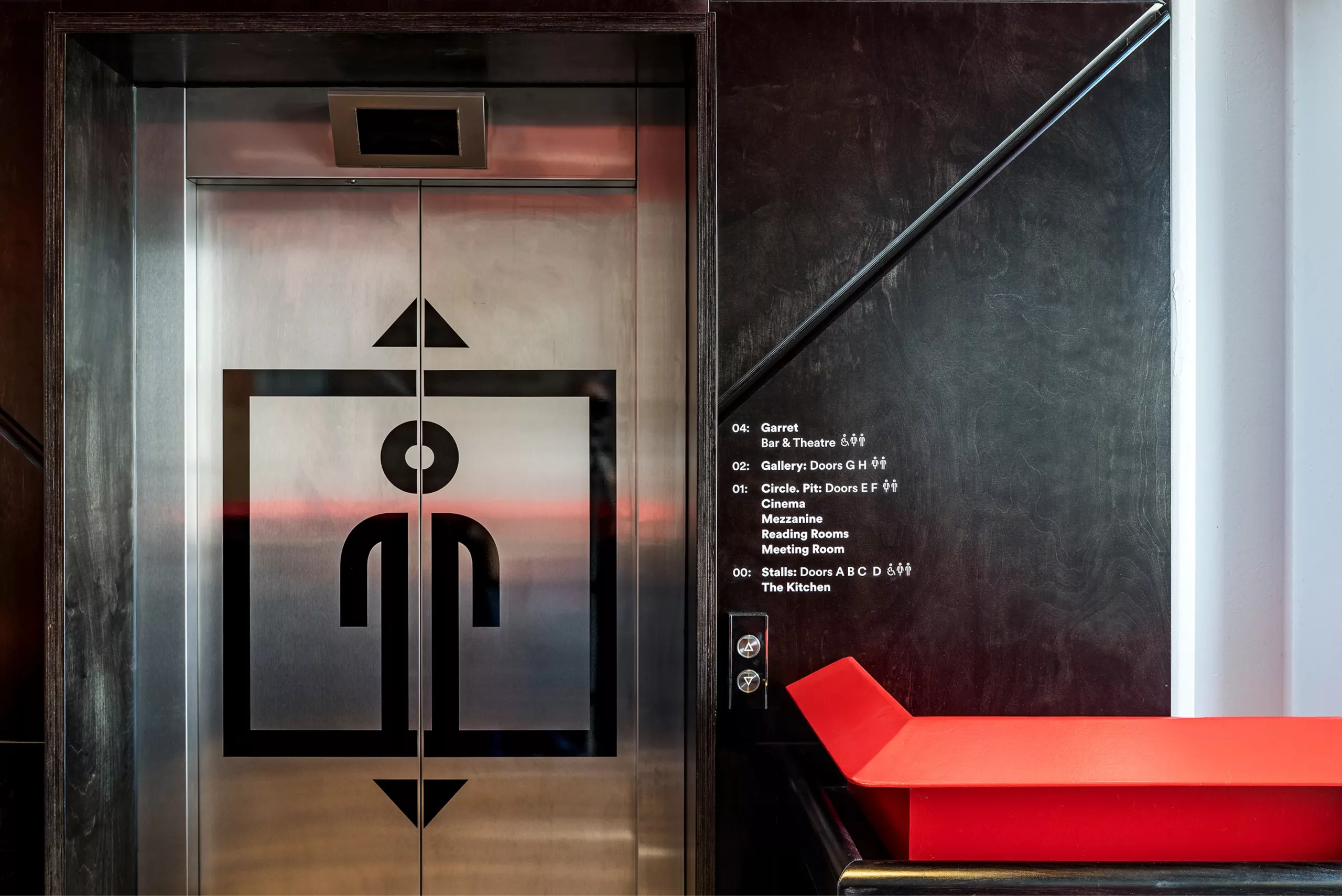 Elevator doors with bold black stylised person icon and up/down arrows, beside a dark wall with white directory text and a red bench.