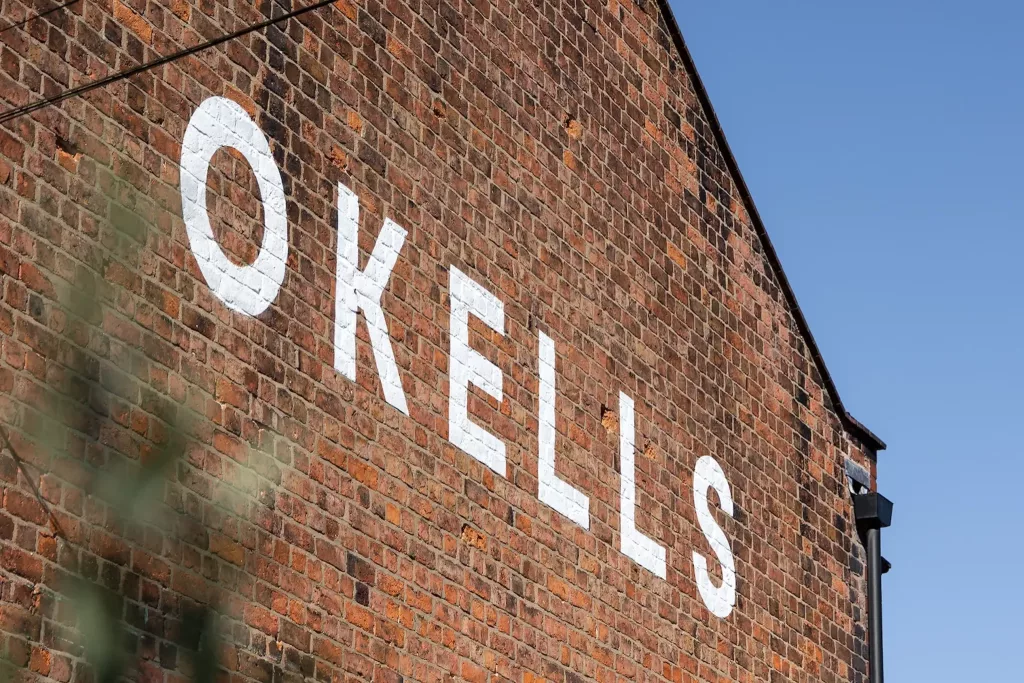 Brick gable wall with large white painted letters reading OKELLS diagonally across it, blue sky and a gutter at the right.