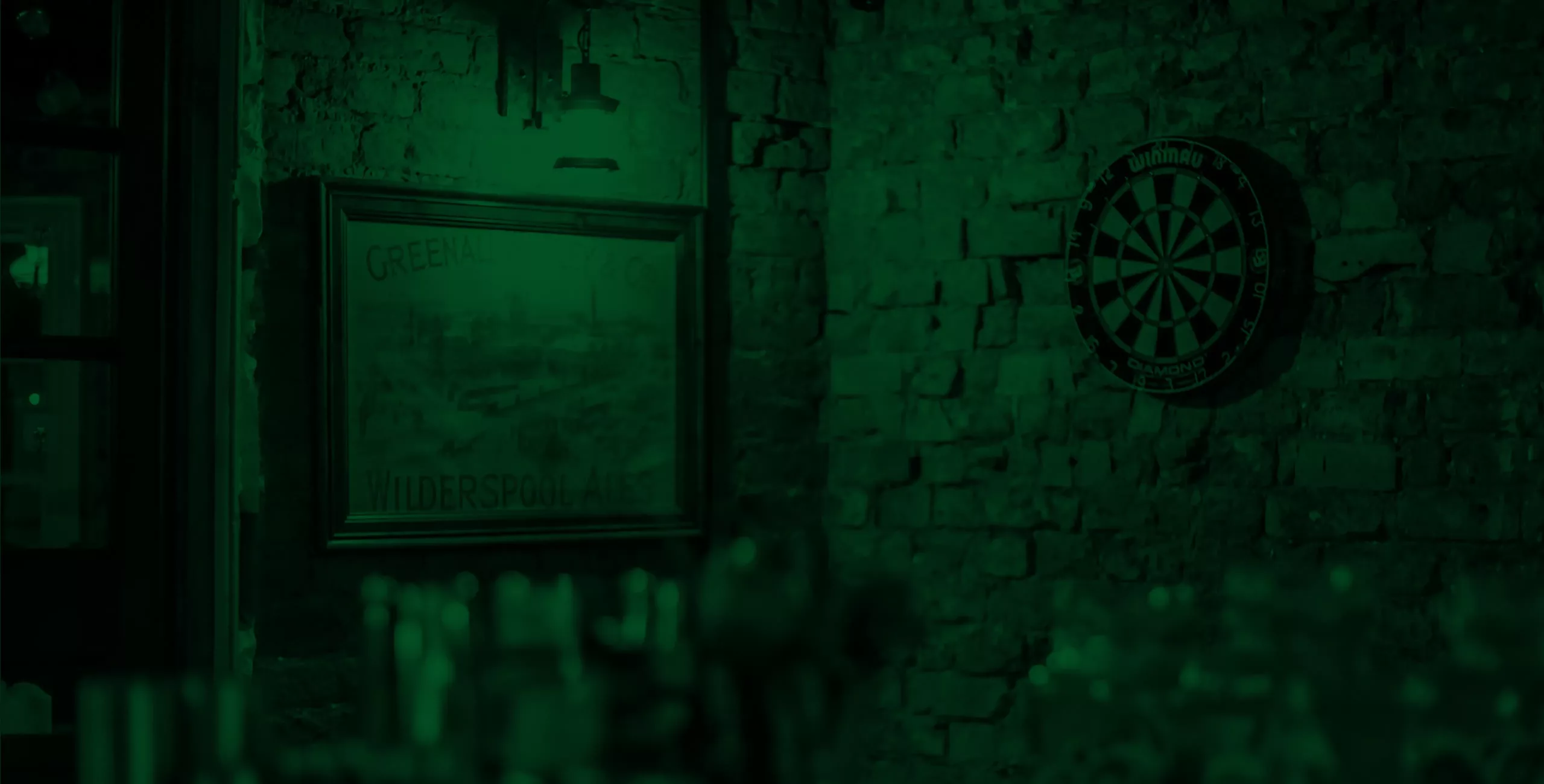 Exposed brick bar wall lit in green with a dartboard, a framed vintage poster and blurred glasses on the bar.