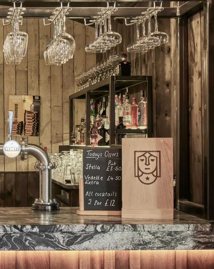 Rustic wooden bar counter with hanging wine glasses, Vedett beer tap, bottles on shelves and chalkboard menu.