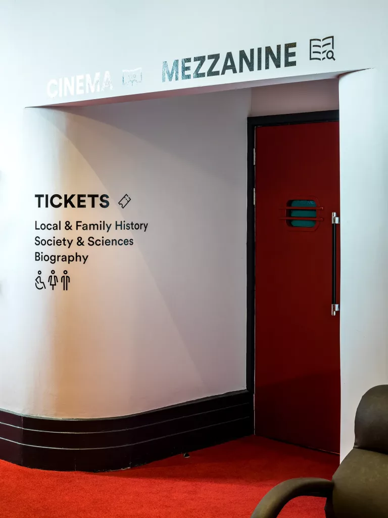 Curved cinema wall with TICKETS list of history, society and biography, accessibility icons and a red door under a MEZZANINE sign.
