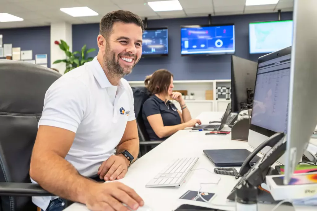Smiling man in white polo using a desktop computer at a bright open-plan office desk, colleague and wall monitors behind.
