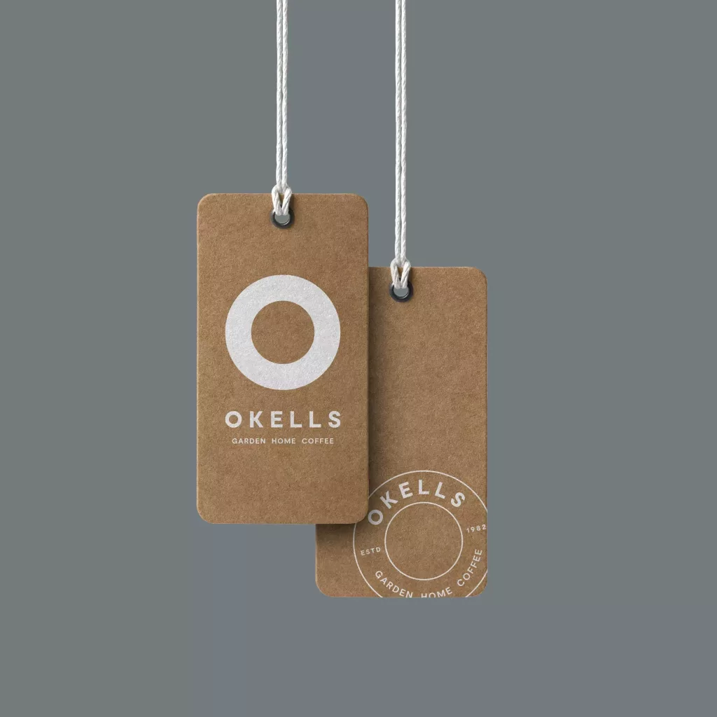 Two brown kraft hang tags suspended by white cords, featuring a white circular logo and the word OKELLS on a grey background.