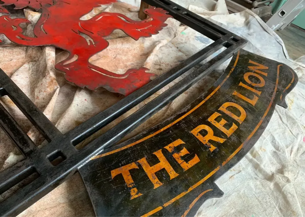 Worn curved pub sign reading THE RED LION in yellow on black with red lion cutout and black frame on a cloth.