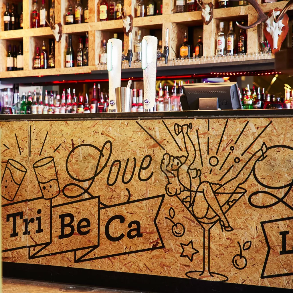 Bar with plywood front reading Love and Tri Be Ca, showing a pin-up in a martini glass and beer taps with bottles behind.