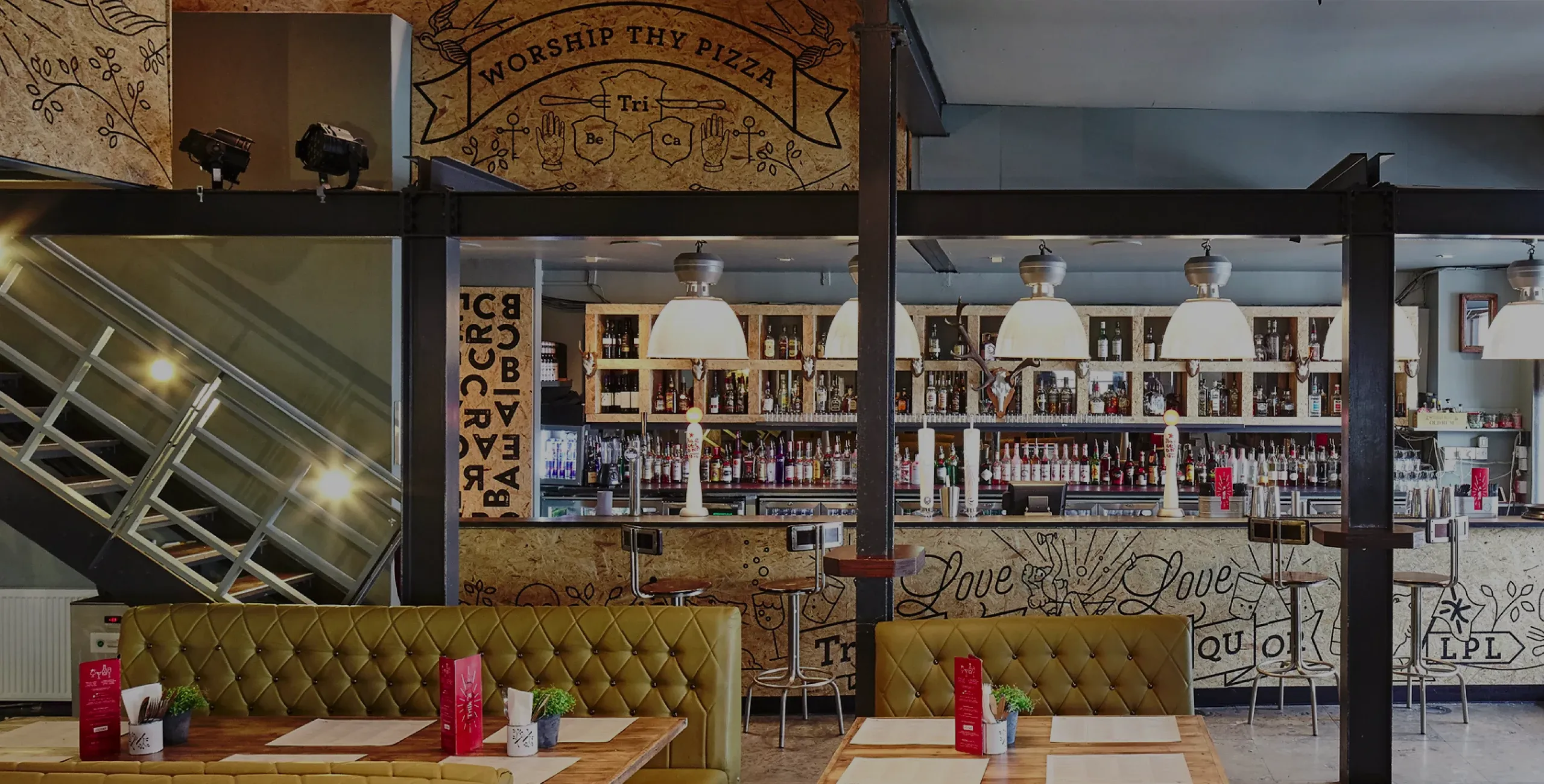 Bar interior with tufted green banquettes, wooden tables with menus, and a back bar lit by pendant lamps and bottles.