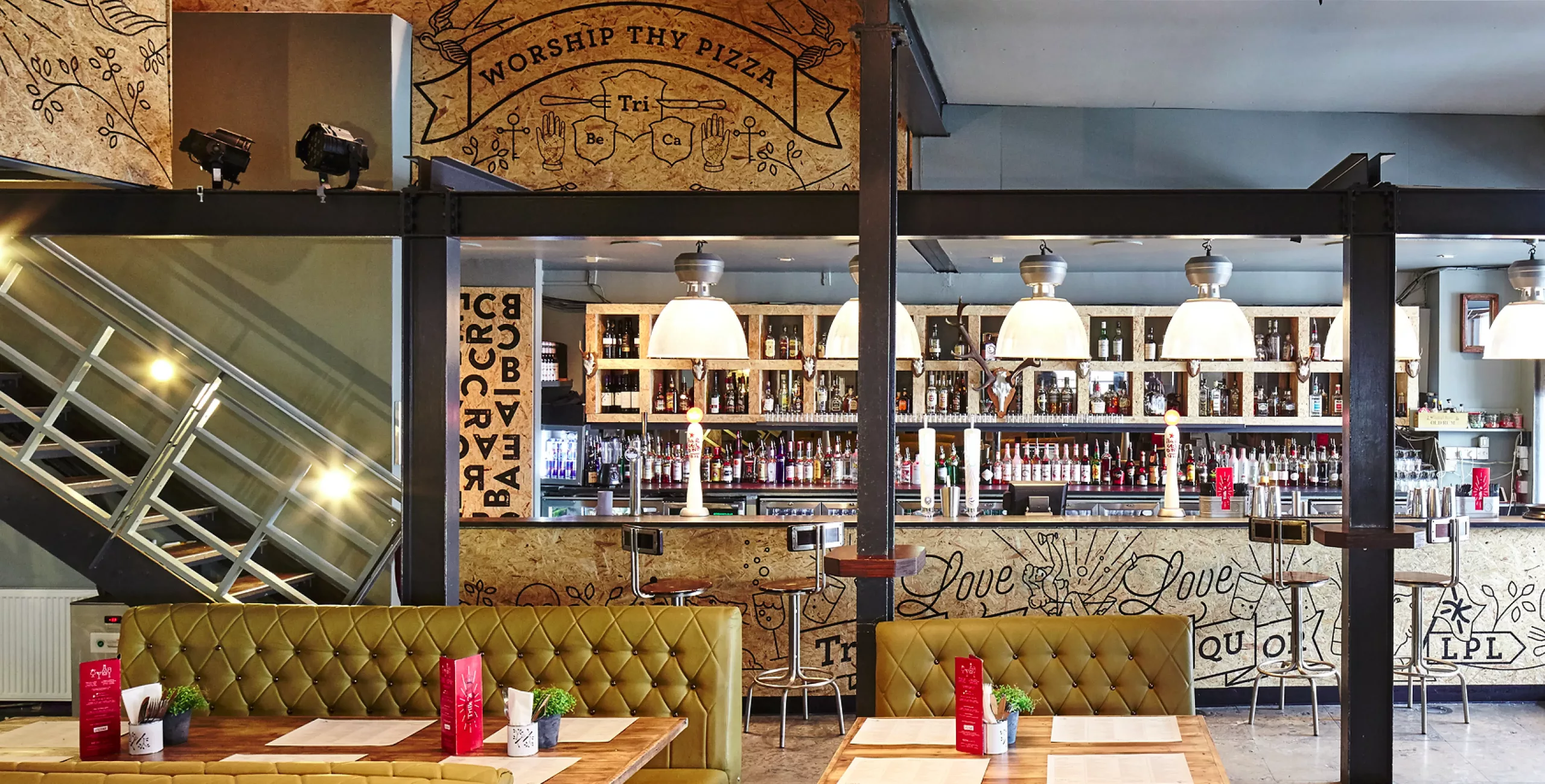 Rustic pizzeria interior with tufted olive-green booths, wooden tables, a long backlit bar with pendant lamps and bottles.