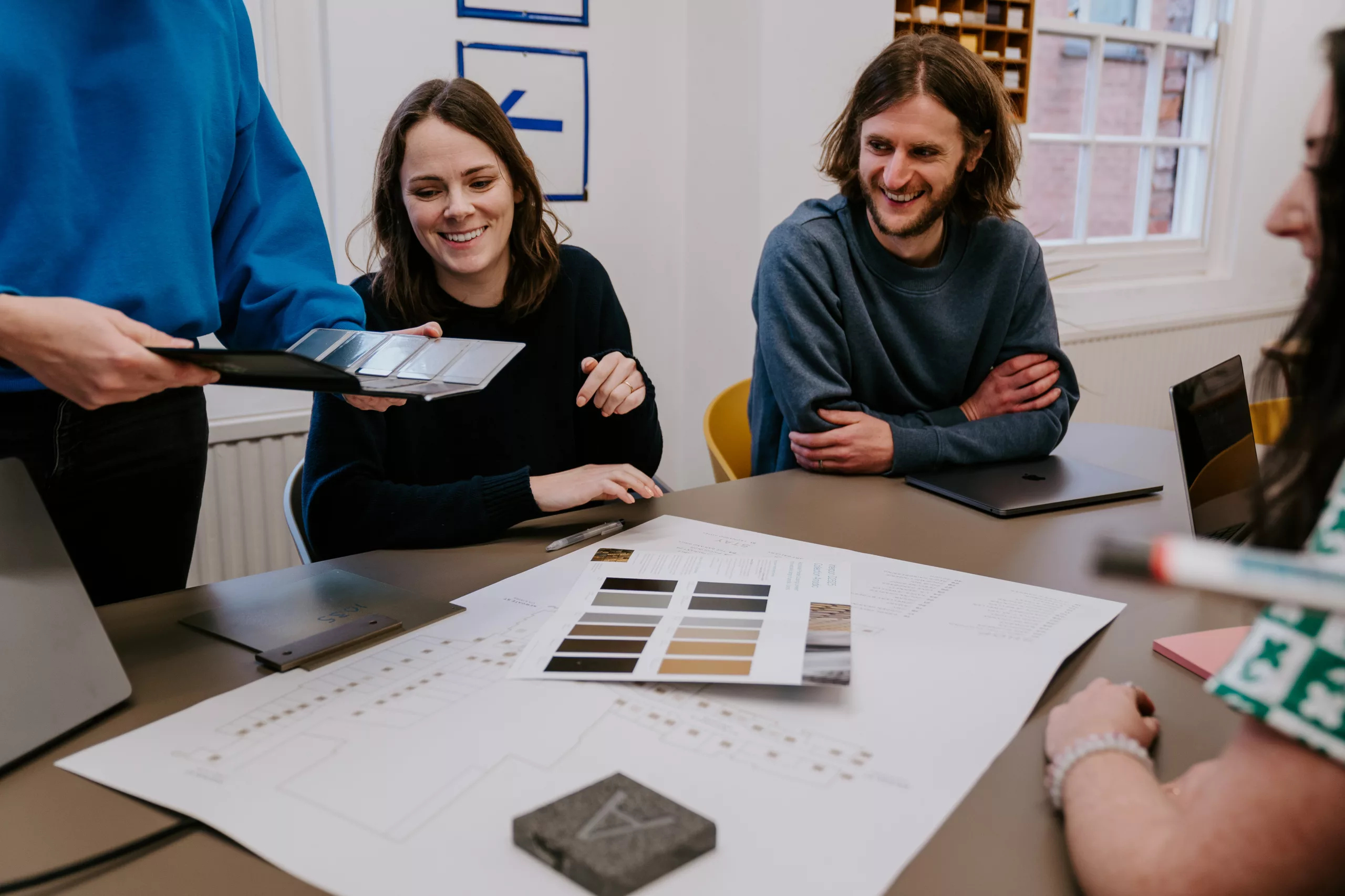 Team smiling over a table of colour swatches, sample tiles, laptops and plans during a design meeting.
