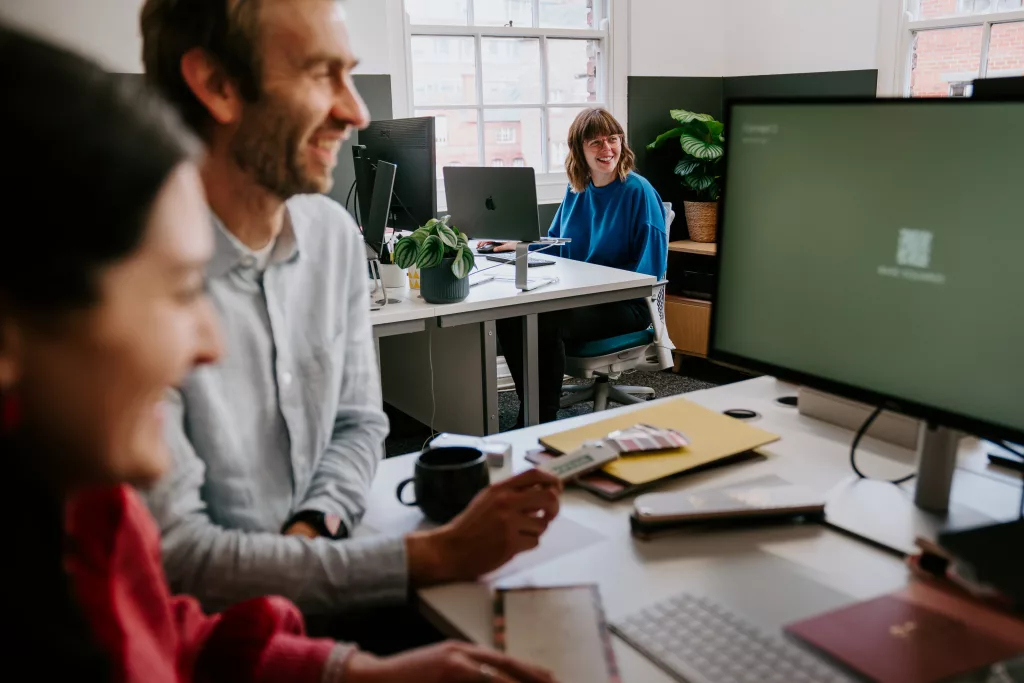 Smiling woman in a blue sweater at a desk with a laptop and plant, two colleagues blurred in the foreground in a bright office.